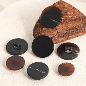 May include: A collection of seven round buttons in varying sizes, featuring smooth surfaces in shades of dark brown and black. Some buttons display subtle, light-colored linear patterns or marbling, while others show the back with a shank attachment.