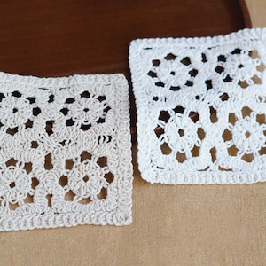 Può includere: Due sottobicchieri quadrati all'uncinetto con motivo floreale. Un sottobicchiere è color bianco sporco e l'altro è bianco brillante. Ogni pezzo è composto da quattro piccole unità quadrate unite tra loro con un bordo decorativo esterno.