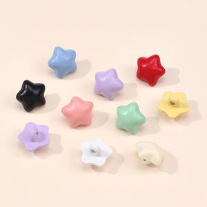Könnte beinhalten: Eine Sammlung von zehn sternförmigen Knöpfen in verschiedenen Unifarben, darunter Hellblau, Schwarz, Lavendel, Rosa, Rot, Minzgrün, Gelb, Weiß und Creme. Jeder Knopf hat eine glatte, abgerundete Oberfläche und eine Öse auf der Rückseite.