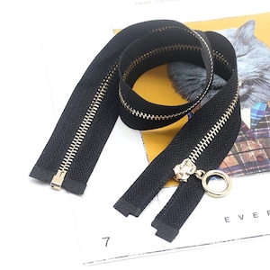 May include: A black zipper with metallic gold-toned teeth and a circular gold-toned pull tab. The zipper is coiled in a loop, showcasing the black fabric tape and the consistent spacing of the metal teeth along the length of the fastener.