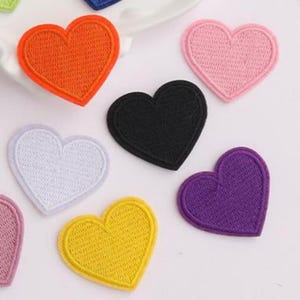 Può includere: Una collezione di toppe ricamate a forma di cuore in vari colori solidi, tra cui arancione, rosa chiaro, nero, bianco, viola e giallo. Ogni toppa presenta una trama cucita densa con un bordo rifinito coordinato a forma di cuore.