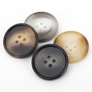 May include: Four round buttons with a recessed center and four holes. The set includes one translucent grey button, one dark brown button with a marbled pattern, one solid black button, and one light tan button with a subtle marbled texture.