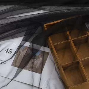 May include: A sheer black tulle fabric draped over a wooden organiser box with six compartments and a white paper sheet featuring the number 45 and a printed photograph of a person in a light-coloured suit, creating a layered visual composition.