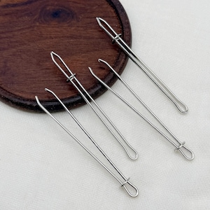 May include: A set of three silver-toned metal bodkin needles, each featuring a long, slender body with a looped end and a sliding ring mechanism designed for threading elastic or ribbon through fabric casings in sewing and craft projects.