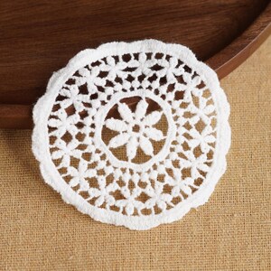 Puede incluir: Un tapete blanco y circular con un patrón de encaje floral. El tapete está hecho de un material suave y texturizado y se coloca sobre una superficie de arpillera beige. El tapete mide aproximadamente 13 cm de diámetro.