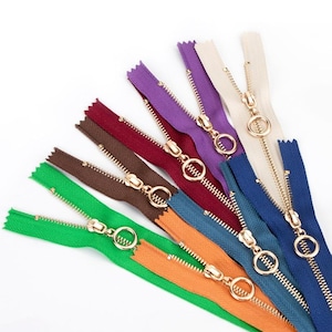 May include: A collection of seven zips in various colours including green, orange, brown, maroon, purple, beige, and blue. Each zip features a gold-toned metal pull tab with a circular ring design and metallic teeth arranged in a fan pattern.