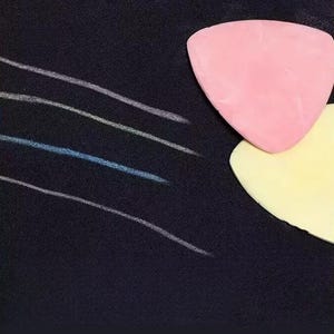 May include: A pink triangular tailor's chalk and a yellow triangular tailor's chalk resting on a dark fabric surface. Four distinct, thin, curved lines in white and light blue are drawn across the black fabric, demonstrating the marking tool.