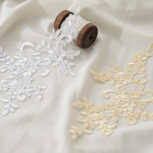 Puede incluir: Apliques de encaje floral blanco y marfil y una bobina de cinta de encaje blanco sobre un fondo de tela blanca. El encaje presenta intrincados diseños florales y de hojas, perfectos para adornar prendas o manualidades.