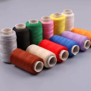 May include: A collection of twelve cylindrical spools of sewing thread arranged in rows. The spools feature a variety of solid colors including grey, black, green, pink, yellow, white, orange, purple, blue, red, beige, and rust brown.