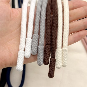 Puede incluir: Una colección de cordones de tela trenzada en blanco, gris claro y marrón oscuro. Cada cordón cuenta con un herrete tejido a juego en el extremo, proporcionando un acabado para sudaderas con capucha, pantalones o prendas ajustables.