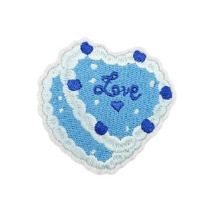 Puede incluir: Un parche bordado en forma de corazón con una base azul claro y bordes festoneados blancos que recuerdan al glaseado. El diseño incluye lunares azul oscuro, puntos blancos pequeños, la palabra Love en cursiva y un pequeño corazón azul.