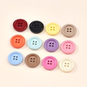 May include: A collection of twelve round, four-hole buttons arranged in three rows. The buttons feature a smooth finish and come in a variety of solid colours including red, black, yellow, tan, orange, light blue, purple, pink, brown, and cream.
