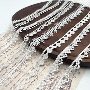 May include: An assortment of white lace ribbons with various delicate patterns, including scalloped edges and floral designs. The ribbons are arranged on a dark wooden surface, showcasing their intricate details. The image captures the texture and elegance of the lace.