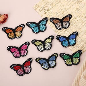 May include: A collection of nine embroidered butterfly patches in various colors including orange, blue, pink, yellow, and green. Each butterfly features black outlines and detailed wing patterns with white spots along the edges of the wings.