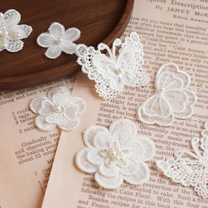 May include: White lace embellishments in various floral and butterfly designs, adorned with small pearl beads. The delicate pieces are arranged on a vintage paper background, evoking a classic, handcrafted aesthetic.