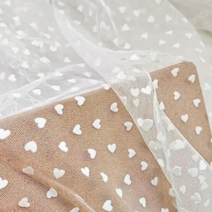 May include: A sheer white tulle fabric featuring a repeating pattern of small, opaque white hearts. The delicate, fine-mesh material is draped in soft folds, creating a layered effect that highlights the texture and the consistent heart design.