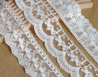 May include: Close-up of three white lace ribbons with intricate floral and geometric patterns. The lace is delicate and detailed, perfect for crafting or embellishing garments. The ribbons are displayed on a wooden surface.