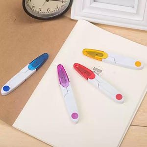 Puede incluir: Cuatro cortahilos plegables con mangos de plástico blanco y tapas de colores translúcidos en azul, morado, rojo y amarillo. Cada herramienta presenta un pequeño acento circular en el mango que coincide con el color de su tapa translúcida.