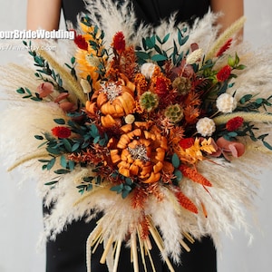 Pode incluir: Um buquê de flores secas com uma mistura de flores laranja, vermelhas e brancas, e capim-dos-pampas bege. O buquê é segurado por uma pessoa que veste um vestido preto.