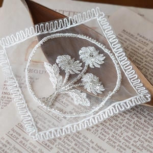 May include: A square white lace patch featuring a central circular motif with embroidered silver floral details. The border consists of a decorative scalloped edge, and the entire piece is crafted from sheer white mesh fabric with intricate stitching.