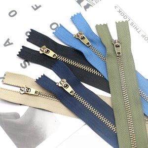 Puede incluir: Un conjunto de cinco cremalleras metálicas en verde oliva, azul claro, negro, azul marino y beige. Cada cremallera cuenta con un cursor metálico y un tirador sujetos a una cinta de tela, dispuestos en forma de abanico sobre una superficie blanca.