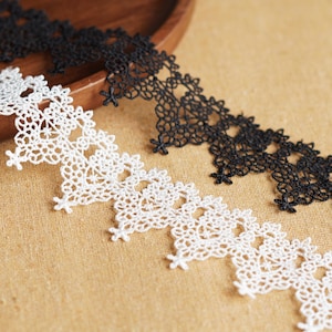 May include: Close-up of two lace trims, one white and one black, with a floral design. The lace is placed on a tan surface, with a wooden bowl in the background. The lace is likely used for sewing or crafting.