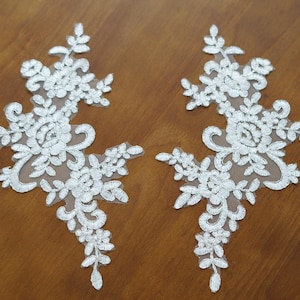 Könnte beinhalten: Zwei weiße Spitzenapplikationen mit floralen und Blattmustern. Die symmetrischen Designs haben ein zentrales Blumenmotiv und filigrane Kanten. Sie liegen auf einer braunen Holzoberfläche.