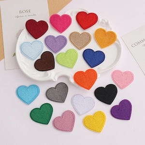 May include: A collection of heart-shaped embroidered patches in a variety of solid colours including red, pink, yellow, blue, green, orange, purple, brown, grey, black, and white, arranged on and around a white scalloped decorative tray.