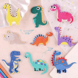 May include: A collection of nine embroidered dinosaur patches in various colours including yellow, green, pink, beige, orange, purple, and blue. Each patch features a cartoon-style dinosaur with distinct patterns, spikes, and spots, arranged on a light surface.