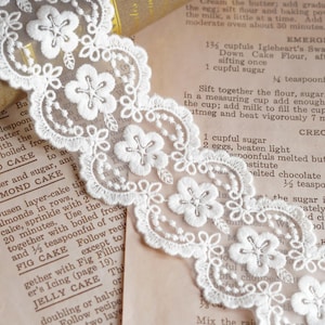 May include: White floral lace trim with a scalloped edge, displayed over vintage recipe pages. The lace features a repeating pattern of stylized flowers and delicate detailing, perfect for crafts and sewing projects.