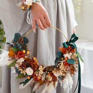Pode incluir: Uma coroa de flores secas com um aro dourado, flores brancas, laranja e marrons, e folhas verdes. Uma fita verde escura está amarrada em torno do aro.