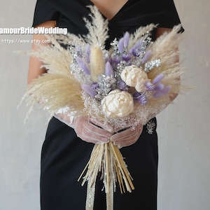 Pode incluir: Um buquê de noiva com capim-dos-pampas, detalhes em lavanda e flores creme. O buquê é amarrado com caules naturais e renda. O texto "GlamourBrideWedding" e um endereço de site são visíveis.