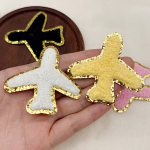 May include: A collection of airplane-shaped patches featuring textured, fuzzy fabric in white, yellow, and pink, each outlined with a shimmering gold sequin border. One black airplane patch with a gold sequin border is visible in the background.