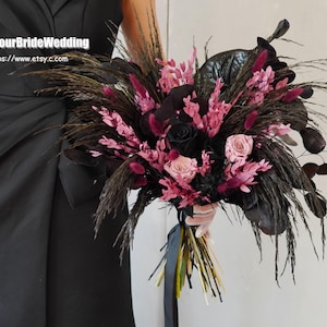 Pode incluir: Um buquê de noiva com penas pretas, flores secas rosa e bordô e folhas pretas. O buquê é amarrado com uma fita preta.