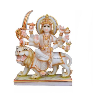 May include: White marble statue of a Hindu goddess riding a lion. The goddess has multiple arms and is adorned with gold accents. The statue is on a marble base.