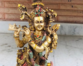 Puede incluir: Una estatua dorada de Krishna tocando la flauta. La estatua está decorada con gemas de colores y detalles intrincados. La estatua está de pie sobre una base con un diseño de loto.