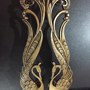 Puede incluir: Un par de manijas de puerta de latón con un diseño de pavo real en espejo. Cada manija muestra un cuello y cabeza alargados en la base, con plumas detalladas que se extienden hacia arriba en un patrón curvo y fluido con un acabado antiguo texturizado.