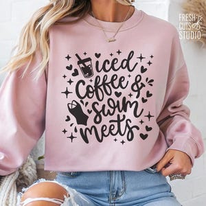 May include: Pink sweatshirt with the text "Iced Coffee & Swim Meets" in black lettering. The text is surrounded by small hearts and stars.