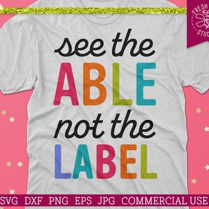 Może przedstawiać: Biały t-shirt z tekstem "see the ABLE not the LABEL" w kolorowych literach. Tekst jest napisany czarnym, ozdobnym pismem. T-shirt leży na różowym tle z złotymi gwiazdkami.