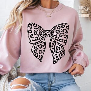 May include: A pink sweatshirt with a black and white leopard print bow design. The sweatshirt has a crew neck and long sleeves.