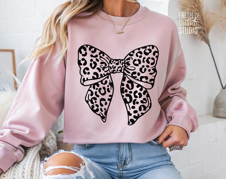 May include: A pink sweatshirt with a black and white leopard print bow design. The sweatshirt has a crew neck and long sleeves.