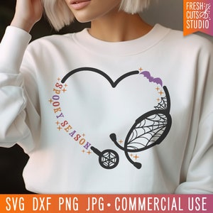 Op de afbeelding: Een wit sweatshirt met een zwart en paars ontwerp van een stethoscoop in de vorm van een hart. De tekst "Spooky Season" staat in kleurrijke letters rond het hart.