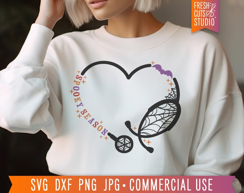 Op de afbeelding: Een wit sweatshirt met een zwart en paars ontwerp van een stethoscoop in de vorm van een hart. De tekst "Spooky Season" staat in kleurrijke letters rond het hart.