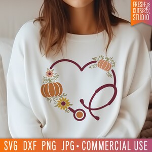Op de afbeelding: Een wit sweatshirt met een bordeauxrood stethoscoopontwerp in de vorm van een hart. De stethoscoop is versierd met herfstthema-elementen, waaronder pompoenen, bladeren en bloemen.