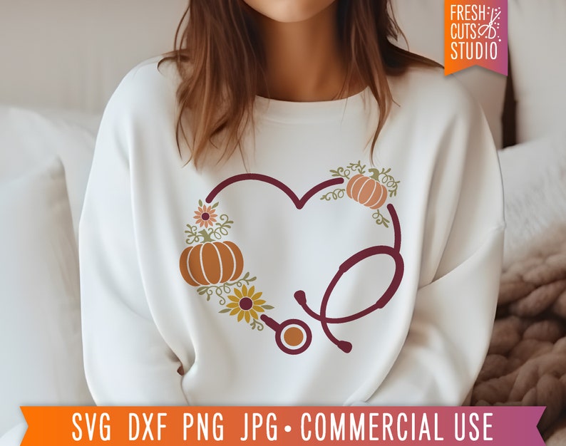 Op de afbeelding: Een wit sweatshirt met een bordeauxrood stethoscoopontwerp in de vorm van een hart. De stethoscoop is versierd met herfstthema-elementen, waaronder pompoenen, bladeren en bloemen.
