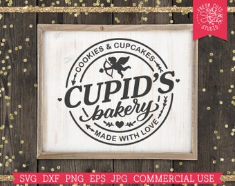Op de afbeelding: Een wit bord met een zwarte cirkel en de tekst "COOKIES & CUPCAKES - CUPID'S bakery - MADE WITH LOVE" in de cirkel. Een silhouet van Cupido met een pijl bevindt zich boven de tekst. Het bord is ingelijst in een lichtbruin houten frame.
