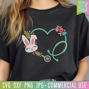 May include: A black t-shirt with a green stethoscope design in the shape of a heart. A pink and white bunny with pink ears is inside the heart. The bunny has pink eyes and a pink nose. The design is surrounded by pink and green flowers and hearts.