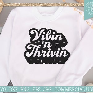 以下が含まれることがあります： 白いスウェットシャツに、レトロなフォントで「Vibin' n Thrivin」という黒い文字が書かれています。文字の周りは小さな白い星で囲まれています。