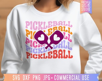 May include: White sweatshirt featuring a colourful graphic design of the word "Pickleball" repeated in a retro font. Two purple pickleball paddles with heart cutouts are centred in the design.