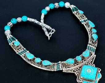 Könnte beinhalten: Eine silberne Halskette mit türkisfarbenen Steinen und Perlen. Die Halskette hat einen zentralen quadratischen türkisfarbenen Anhänger mit floralem Design, umgeben von kleineren tropfenförmigen türkisfarbenen Steinen. Die Halskette hat eine Perlenkette und einen Verschluss.